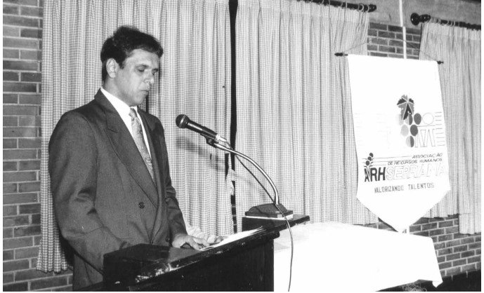 Palestrante ao microfone diante do banner da ARH Serrana, em foto em preto e branco